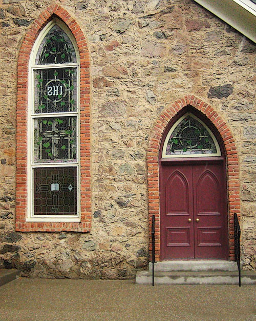 A charming stone Gothic church with burgundy doors and a stained glass window under brick arches.