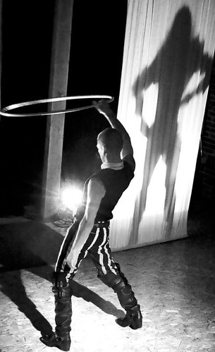 Black and white photo of a person with a shaved head, wearing a white shirt with a black vest, tight black-and-white striped pants, and knee-high black leather boots. They are holding a hula hoop over their head, while the shadow of an apparently long-haired person is cast on a white curtain near them.