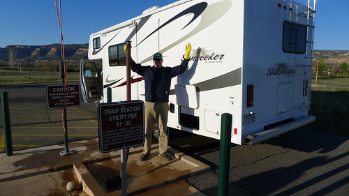 A person standing on the left side of a motorhome with their arms raised, in a lot next to an RV waste disposal station