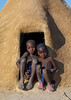 Mucubal tribe children sitting at the entrance of their hut covered of adobe, Namibe Province, Virei, Angola